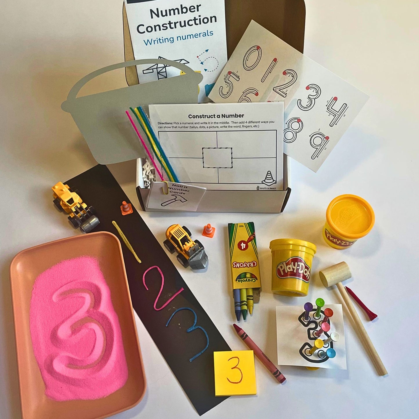 Math in Reach construction-themed monthly math kit for preschool and kindergarten. Includes sand for number tracing, Play-Doh, toy trucks, crayons, wax sticks, and supporting materials to practice writing numerals 0–9.