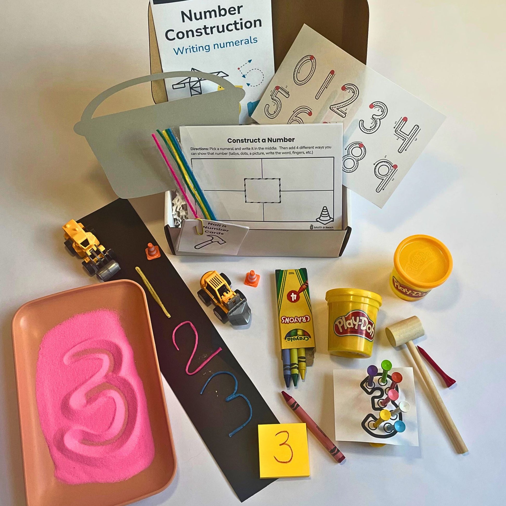 Math in Reach construction-themed monthly math kit for preschool and kindergarten. Includes sand for number tracing, Play-Doh, toy trucks, crayons, wax sticks, and supporting materials to practice writing numerals 0–9.