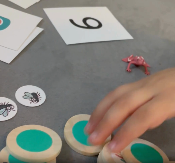 Video of a child using Math in Reach frog-themed counting activity. Shows hands-on play with frog figurines, wooden counting chips, flies, and number cards to practice early math skills for ages 4–6.